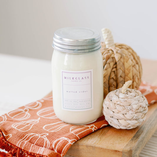 A white soy wax candle in a clear glass jar with a label, placed on an orange cloth with pumpkin patterns, next to a decorative pine cone.